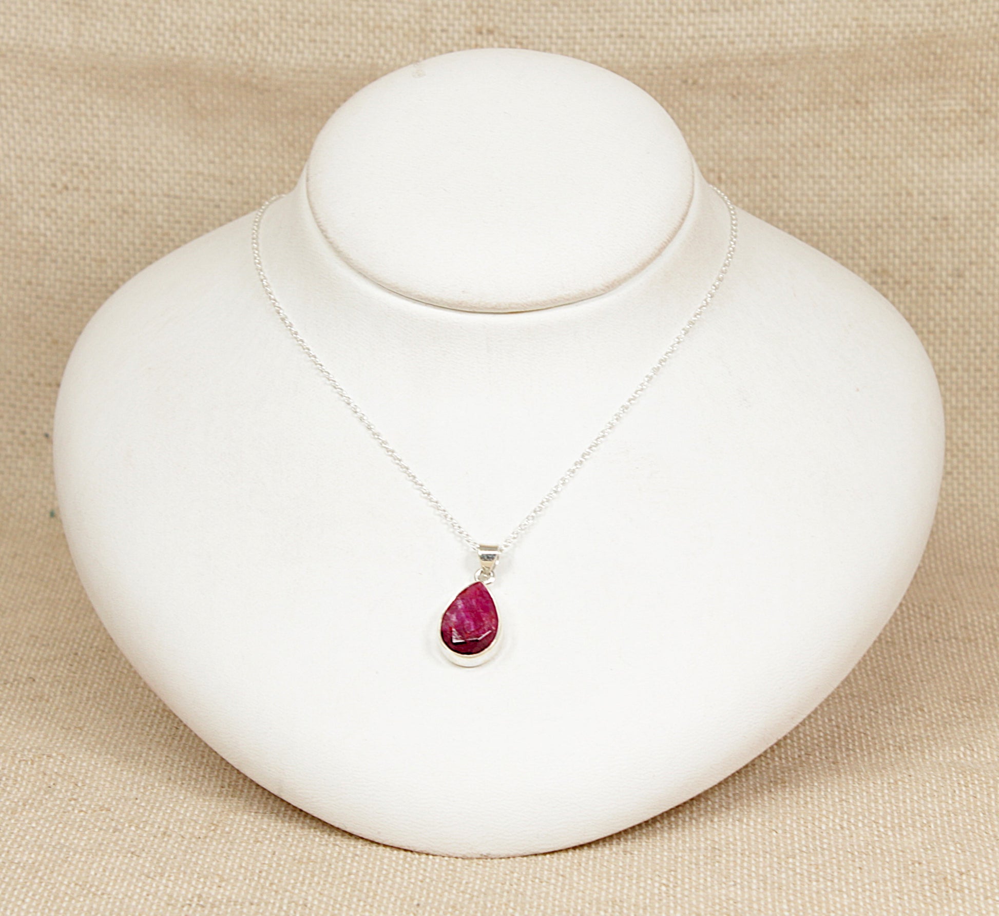 Silver necklace with a teardrop-shaped ruby pendant on a white stand against a beige background