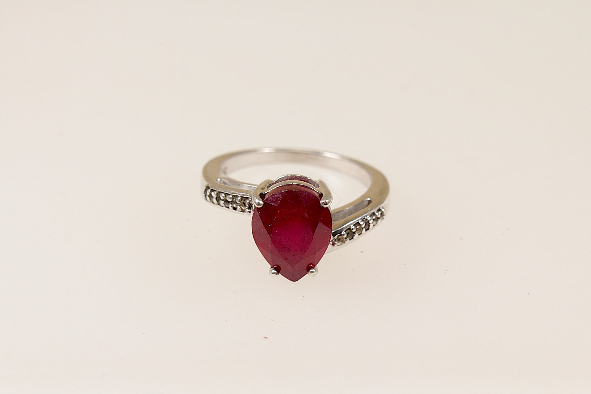 A sterling silver pear shaped ruby solitaire with white topaz accents on each shoulder, Stamped 925 for sterling silver and the makers mark ‘TGGC’ for The Genuine Gemstone Company, UK finger size J.