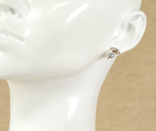 A pair of silver stud earrings in the shape of a dolphin with a round blue topaz placed on a mannequin ear.