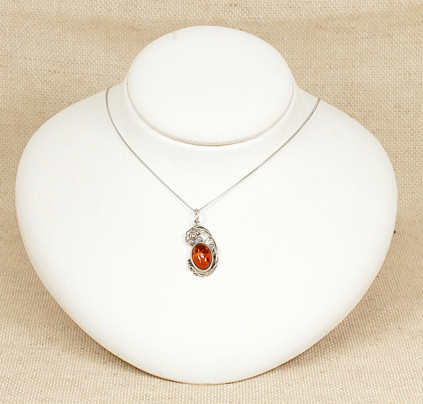 A beautifully crafted pendant featuring an oval piece of genuine Baltic amber, richly golden with delicate inclusions of ancient vegetation suspended within. The amber is bezel-set in finely detailed sterling silver, adorned with ornate floral motifs that echo the graceful curves of the Art Nouveau style. Expertly made and stamped 925 for sterling silver, this timeless piece hangs from an elegant 18-inch silver curb chain.