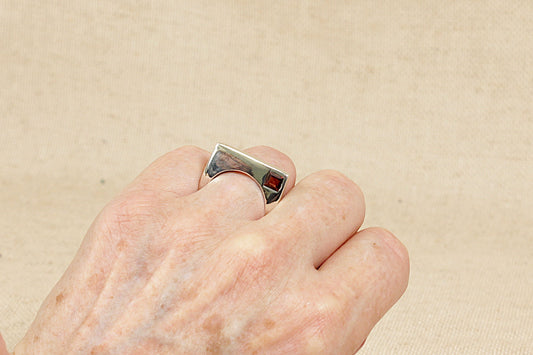 An Interesting Silver and Garnet Modernist Designer Ring.
A striking and contemporary designer sterling silver ring, featuring a unique curved square silhouette with a central round finger cutout for a sculptural, modernist feel. In one corner of the square, a rich square-cut faceted garnet is inset into the silver, adding a bold pop of deep red and a geometric contrast to the fluid shape of the ring.
Minimal yet eye-catching, UK finger size O.