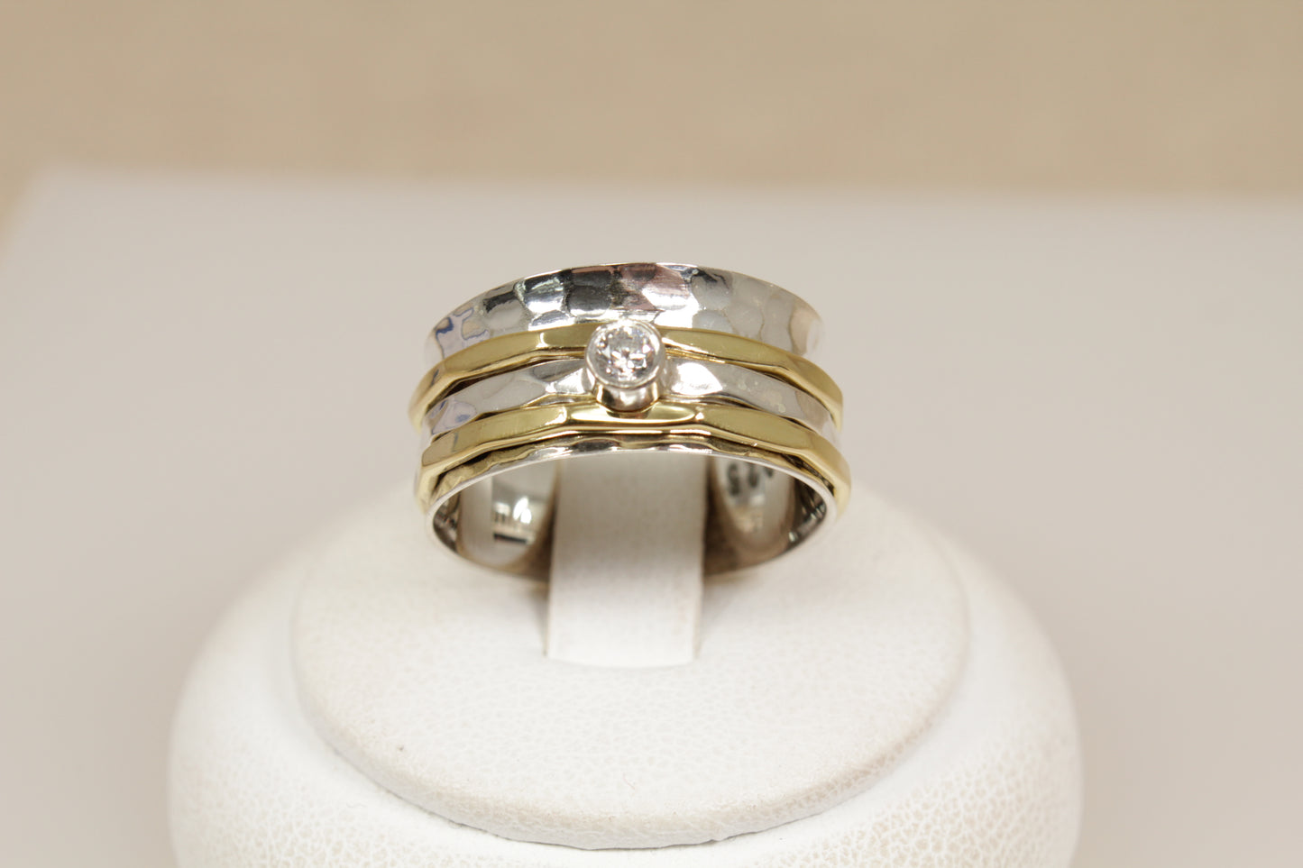 A stylish new Silver spinner ring, two gold colour bands and one silver with white gemstone on a white stand with a beige background. 