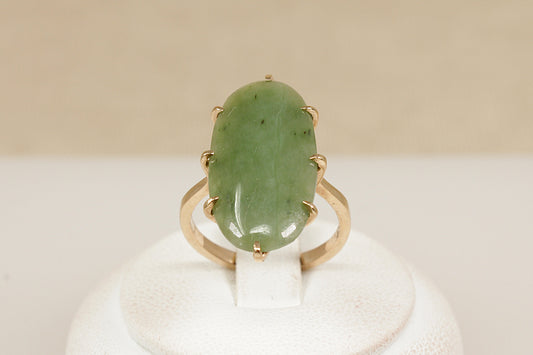A graceful vintage ring crafted in solid 9ct yellow gold, showcasing a striking navette-shaped Jadeite cabochon. The smooth, polished surface of the Jadeite is gently domed, with natural inclusions visible—typical of authentic, untreated jadeite and adding to its organic charm. The inside of the band is stamped ‘9ct’, UK finger size N.