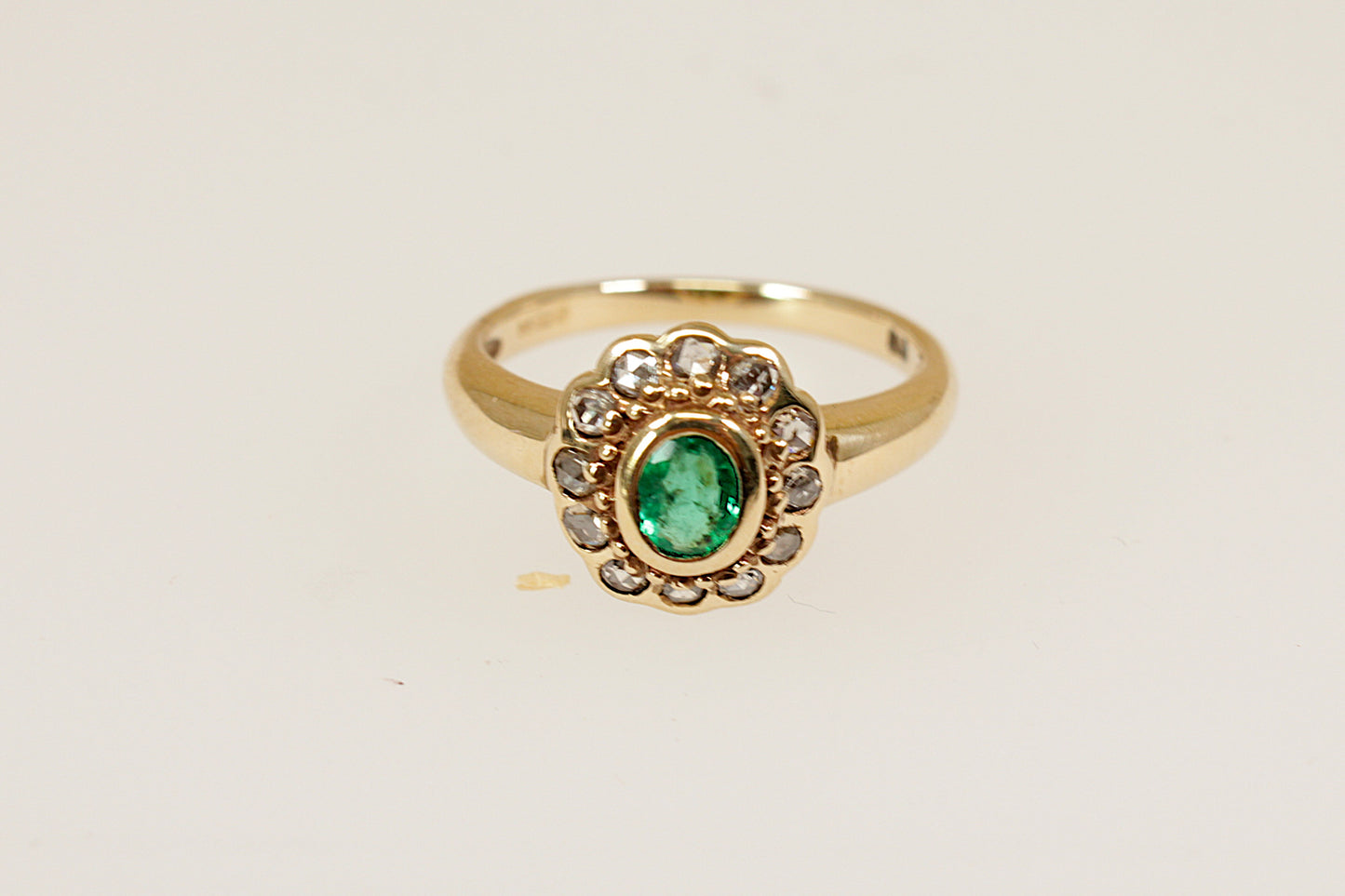 A beautiful vintage cluster ring crafted in 9ct yellow gold, featuring a vibrant oval faceted emerald in a bezel setting. The emerald is slightly raised at the centre of the head, drawing the eye with its rich green hue. Surrounding the central stone are twelve sparkling old-cut diamonds. UK finger size M.