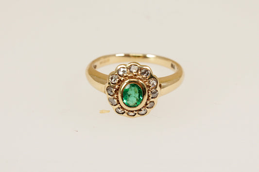 A beautiful vintage cluster ring crafted in 9ct yellow gold, featuring a vibrant oval faceted emerald in a bezel setting. The emerald is slightly raised at the centre of the head, drawing the eye with its rich green hue. Surrounding the central stone are twelve sparkling old-cut diamonds. UK finger size M.