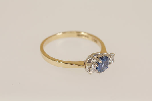 9ct Gold Sapphire and Diamond Trilogy Ring.A timeless and graceful 9ct Gold trilogy ring, featuring a stunning 0.50ct cornflower blue round Sapphire at its centre, securely 4-claw set to allow maximum light and brilliance. Flanking the sapphire are two 0.05ct round-cut Diamonds, the ring is fully hallmarked in Birmingham for 1989, confirming its gold purity and vintage heritage. A classic and romantic piece—perfect for an engagement or anniversary, UK Finger size: M.