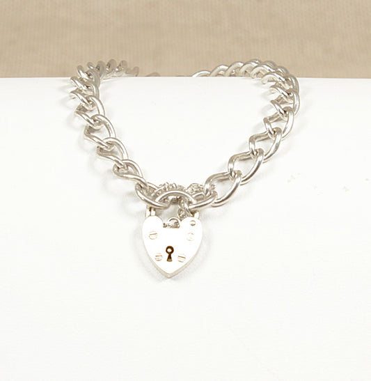 A sterling silver curb link bracelet with a heart-shaped padlock clasp and a safety chain, displayed against a beige background.