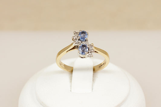 A beautifully designed 9ct gold ring, featuring two oval blue topaz gemstones set vertically with a slight twist, creating a fluid and elegant silhouette. Surrounding the topaz stones are eight sparkling diamonds, gracefully arranged in a flowing S-shape, adding a delicate brilliance and movement to the design, London 1992, UK finger size M.