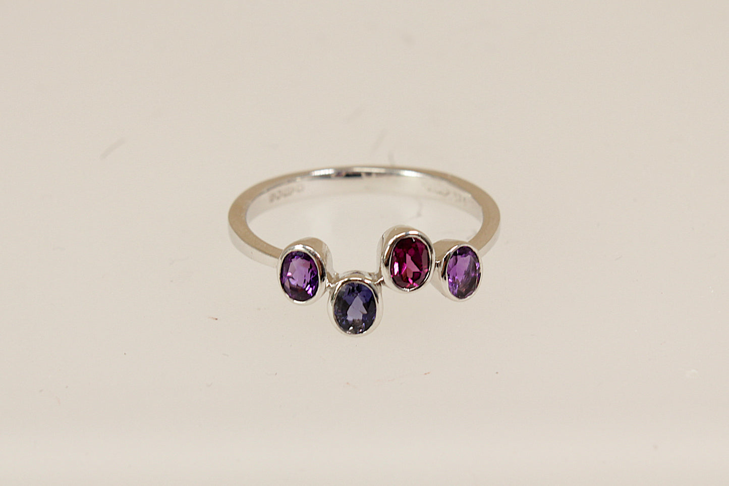 9ct White Gold Multi Gemstone Ring, A delicate and modern 9ct White Gold ring, featuring four oval bezel-set gemstones arranged in a stylish, irregular line across the band. The design includes two vibrant Amethysts, a deep Blue Iolite, and a rich Garnet, full London hallmarks for 2005, UK Finger size: O.
