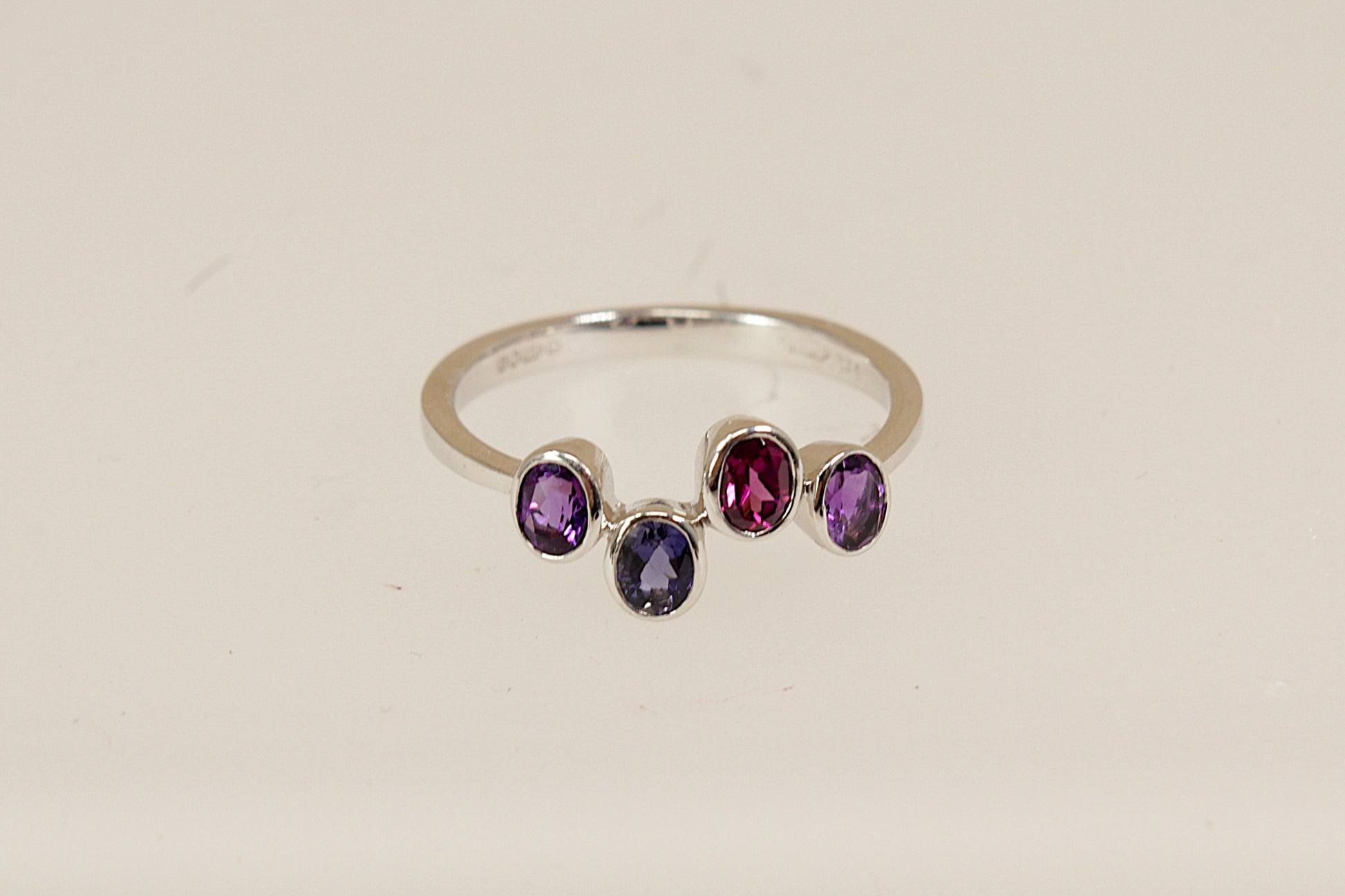 9ct White Gold Multi Gemstone Ring, A delicate and modern 9ct White Gold ring, featuring four oval bezel-set gemstones arranged in a stylish, irregular line across the band. The design includes two vibrant Amethysts, a deep Blue Iolite, and a rich Garnet, full London hallmarks for 2005, UK Finger size: O.